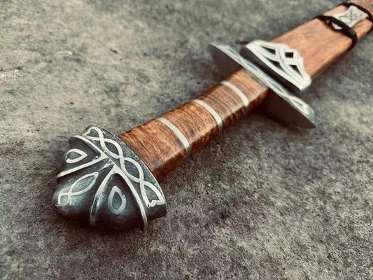 Close-up of the sword pommel and wrapped wooden handle with natural material grip, showing decorative Norse-style metalwork and craftsmanship details.