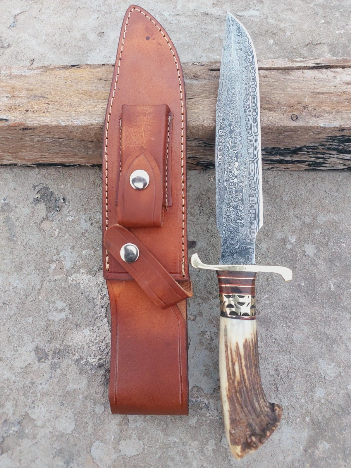 Damascus steel hunting knife with crown stag handle and brass spacers, showcasing traditional craftsmanship. A polished, sharp blade paired with a stitched leather sheath ensures safe storage and portability.