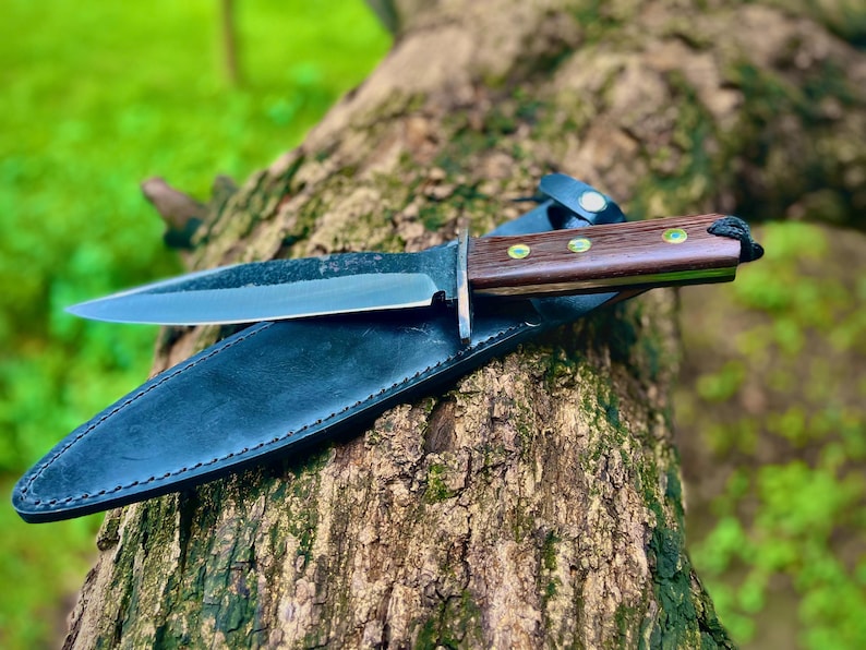 Outdoor photo of carbon steel hunting knife on black leather sheath with polished wooden handle and lanyard.