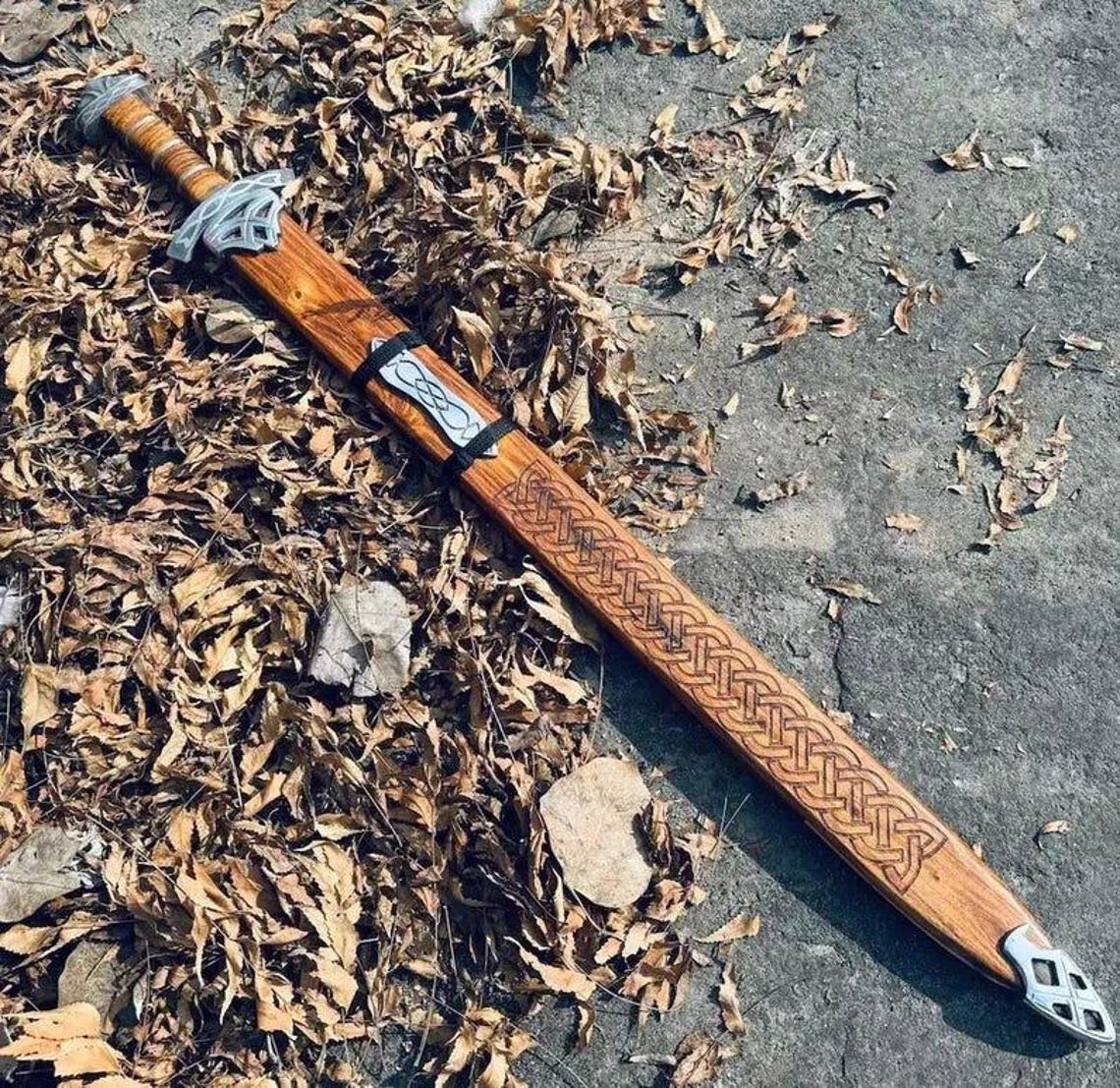 Full view of carved wooden scabbard placed among dry leaves, highlighting detailed Celtic knotwork engraving and the scabbard’s wood grain.