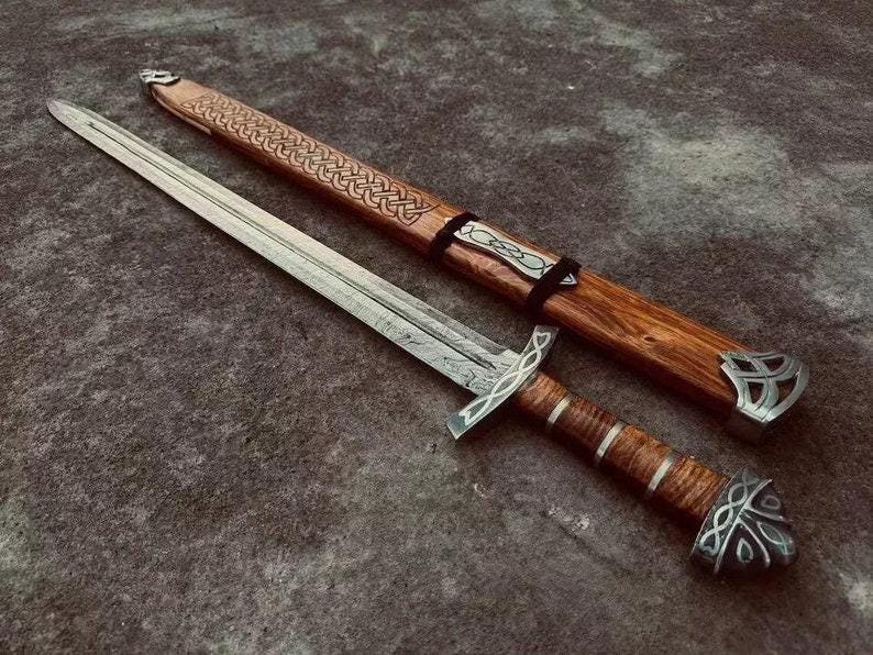 Handmade Viking sword laid beside its scabbard showing full 36" length, patterned Damascus blade, carved wooden scabbard with Celtic knotwork, and metal fittings.