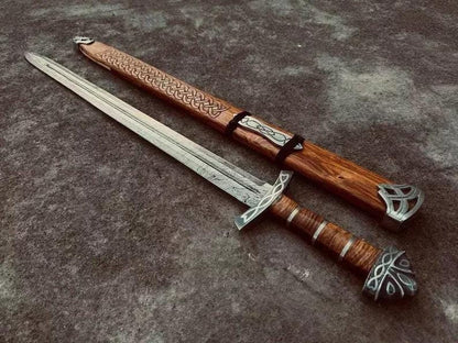 Handmade Viking sword laid beside its scabbard showing full 36" length, patterned Damascus blade, carved wooden scabbard with Celtic knotwork, and metal fittings.