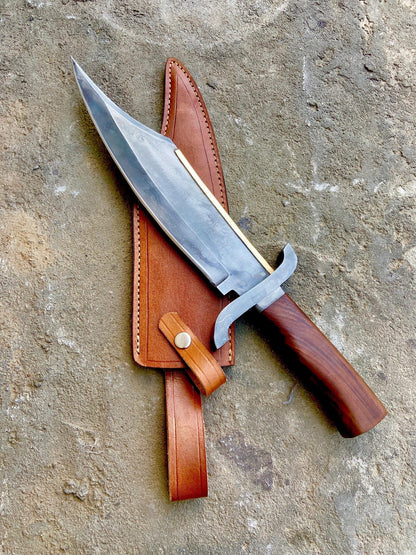 Handmade Alamo Musso bowie knife with 5160 spring steel blade and wooden handle displayed on leather sheath on stone background.