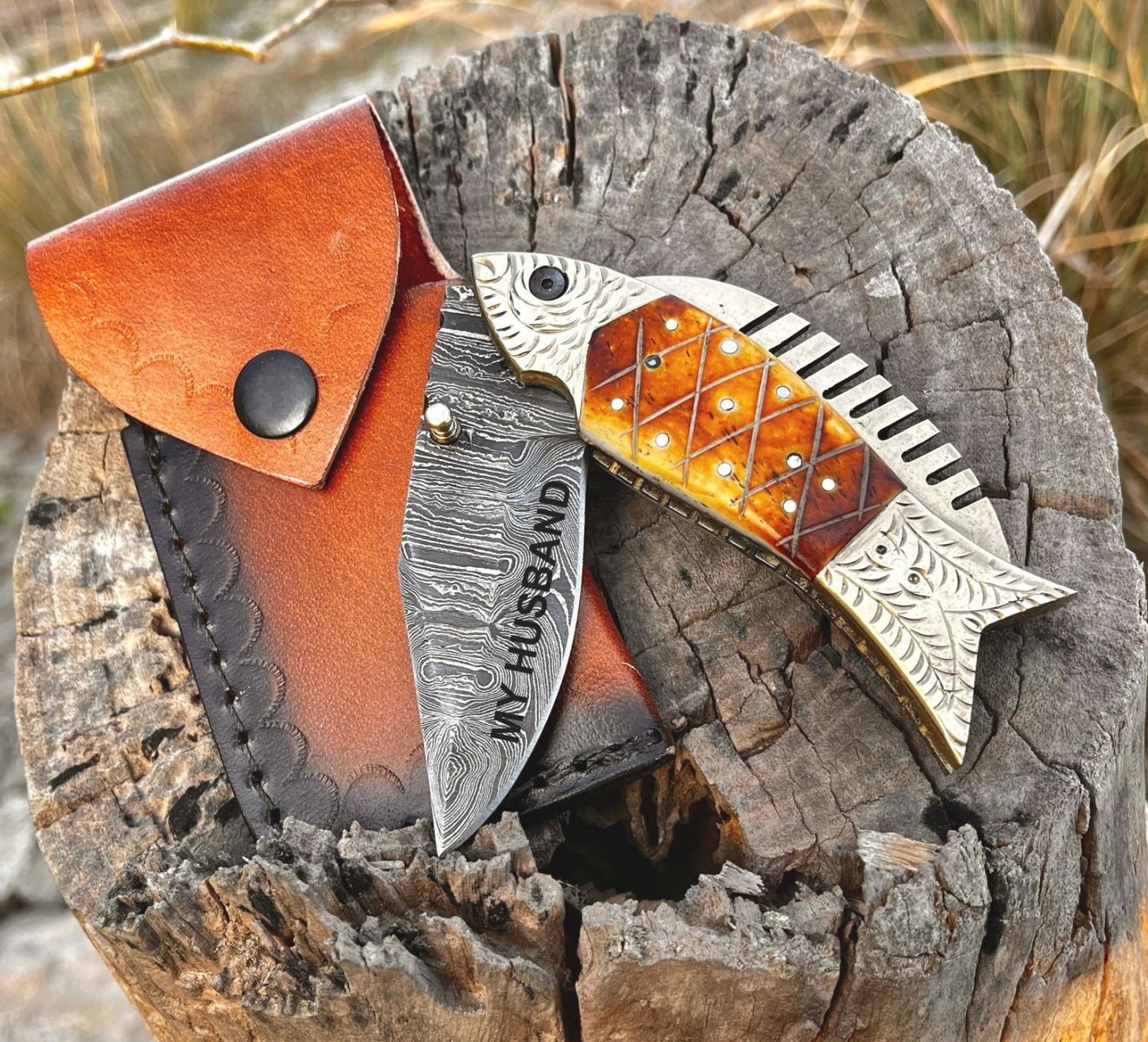 Fish shaped folding knife with personalization and monogram options displayed with leather sheath on wooden background.