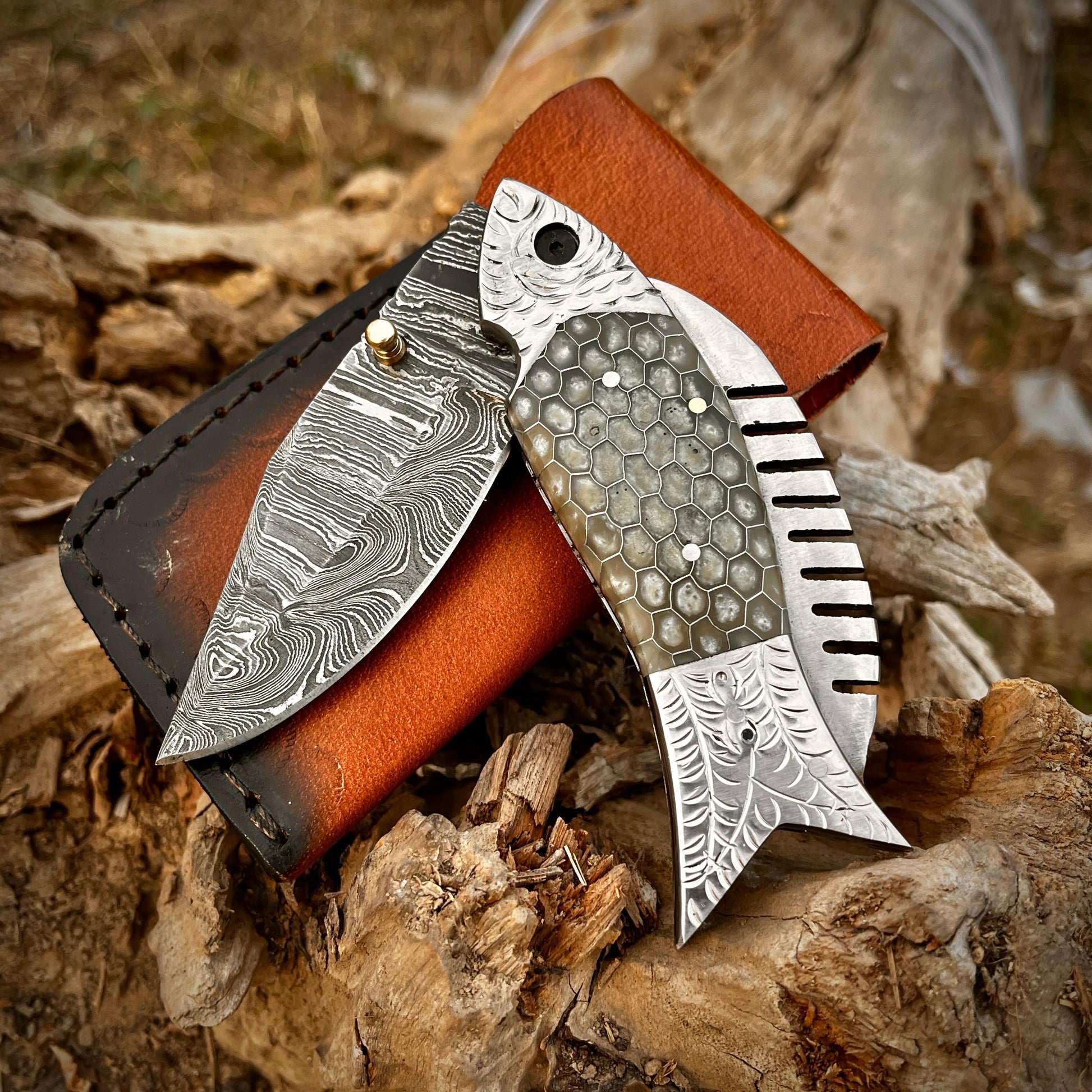 Grey honeycomb fish Damascus folding pocket knife with leather sheath displayed on natural wood background showing detailed Damascus blade pattern and artistic fish shaped handle.