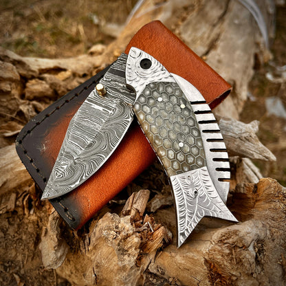 Grey honeycomb fish Damascus folding pocket knife with leather sheath displayed on natural wood background showing detailed Damascus blade pattern and artistic fish shaped handle.