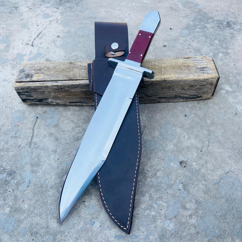 Handmade carbon steel bowie knife with Micarta handle and leather sheath displayed outdoors.