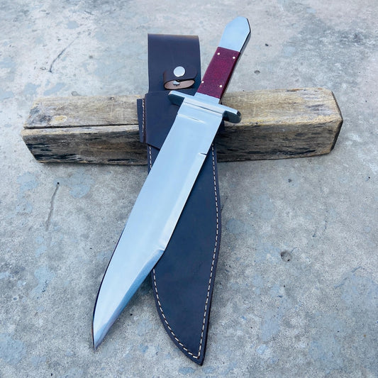 Handmade carbon steel bowie knife with Micarta handle and leather sheath displayed outdoors.