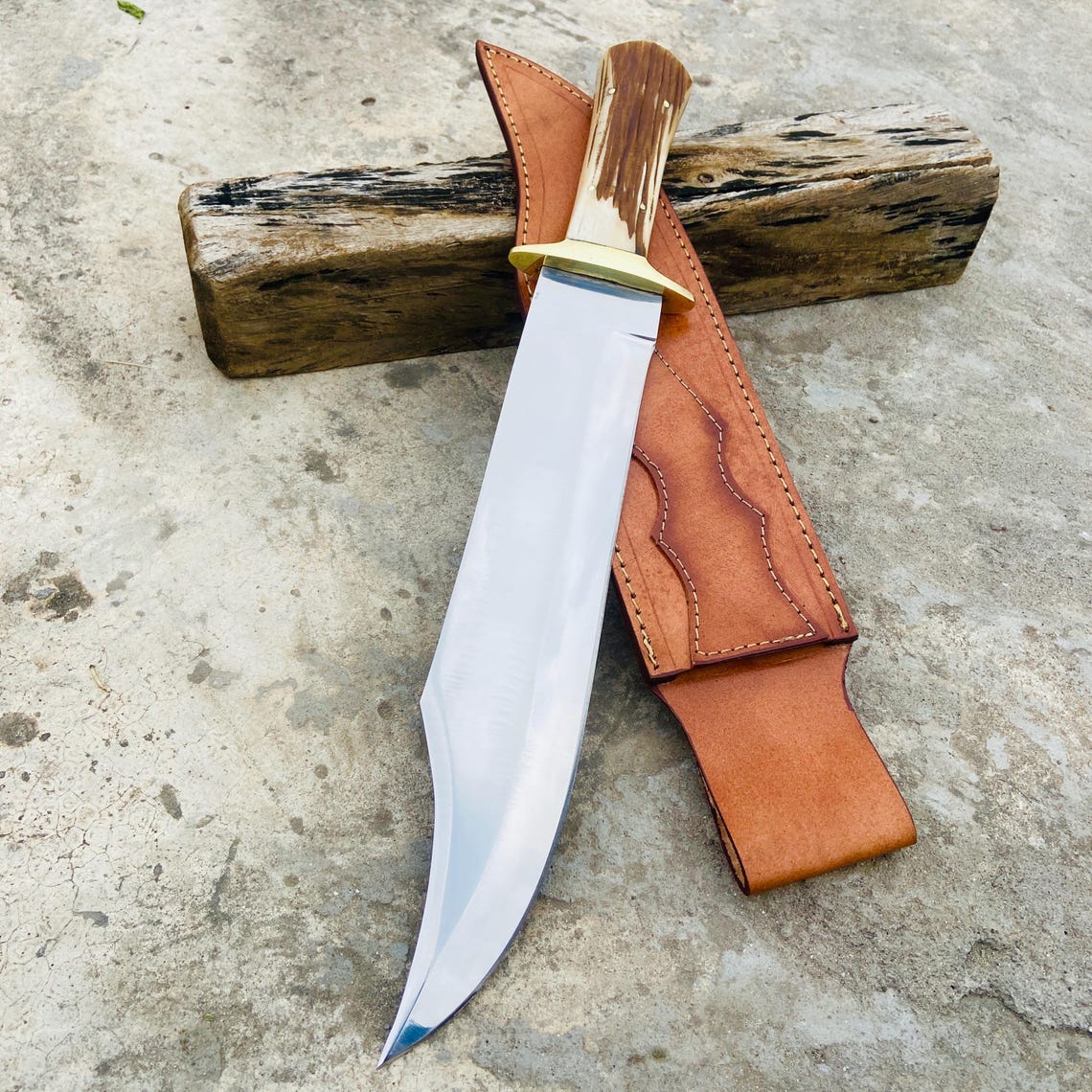 Handmade D2 steel bowie knife with mirror-polished blade, stag handle, and leather sheath displayed on wood.