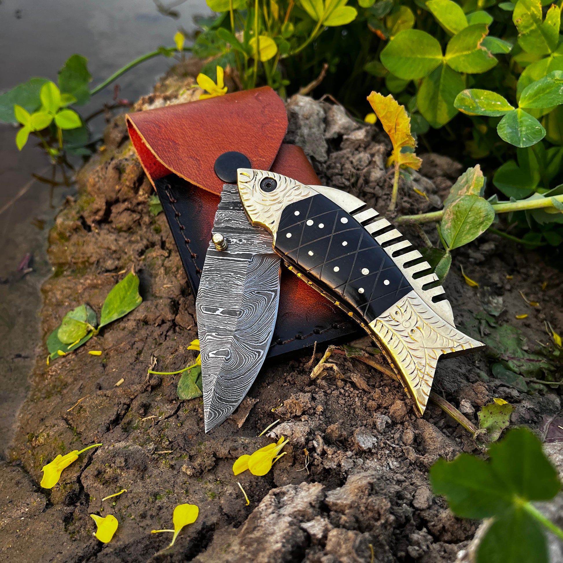 Handmade black scale fish Damascus pocket knife with leather sheath displayed on natural outdoor ground showing detailed Damascus blade pattern and fish shaped handle design.