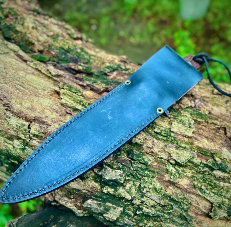 Full black leather sheath for handmade carbon steel hunting knife resting on a tree log.