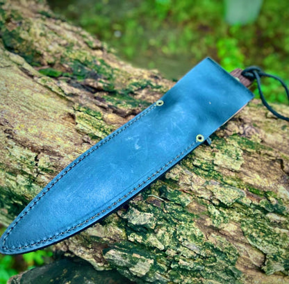 Full black leather sheath for handmade carbon steel hunting knife resting on a tree log.