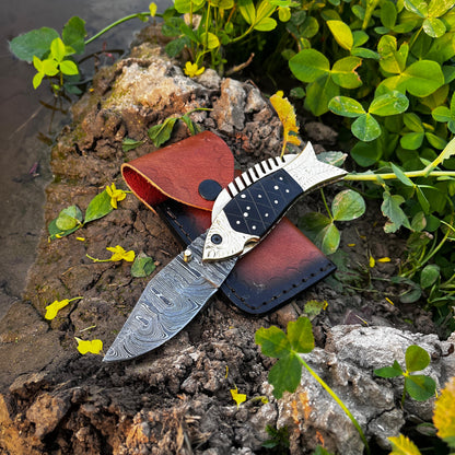 Black scale fish folding knife placed on leather sheath in outdoor setting highlighting Damascus blade texture and unique fish handle craftsmanship.