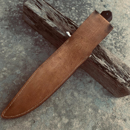 Artisan handmade Bowie knife with carbon steel blade, dark wood handle, and leather sheath arranged on wood.