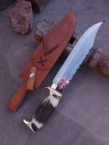 Large handmade Bowie knife with mirror-polished carbon steel blade and leather sheath on rustic surface.