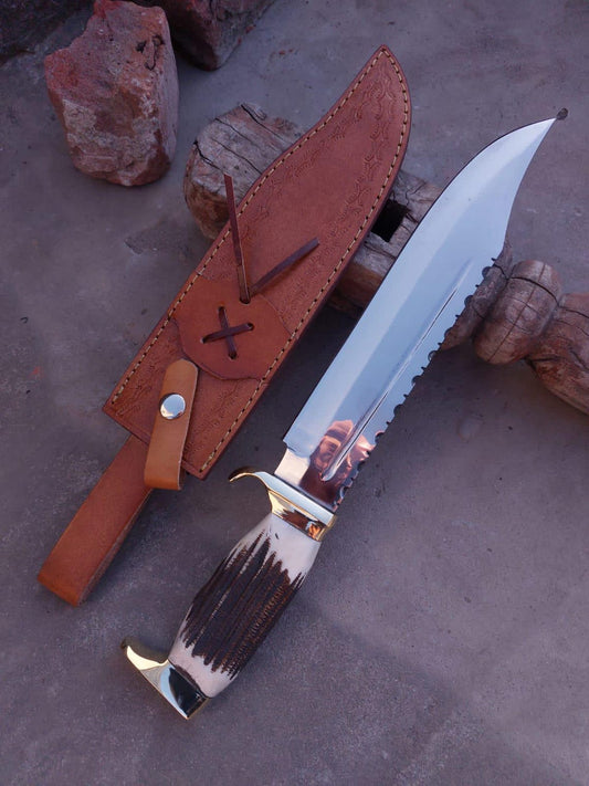 Large handmade Bowie knife with mirror-polished carbon steel blade and leather sheath on rustic surface.