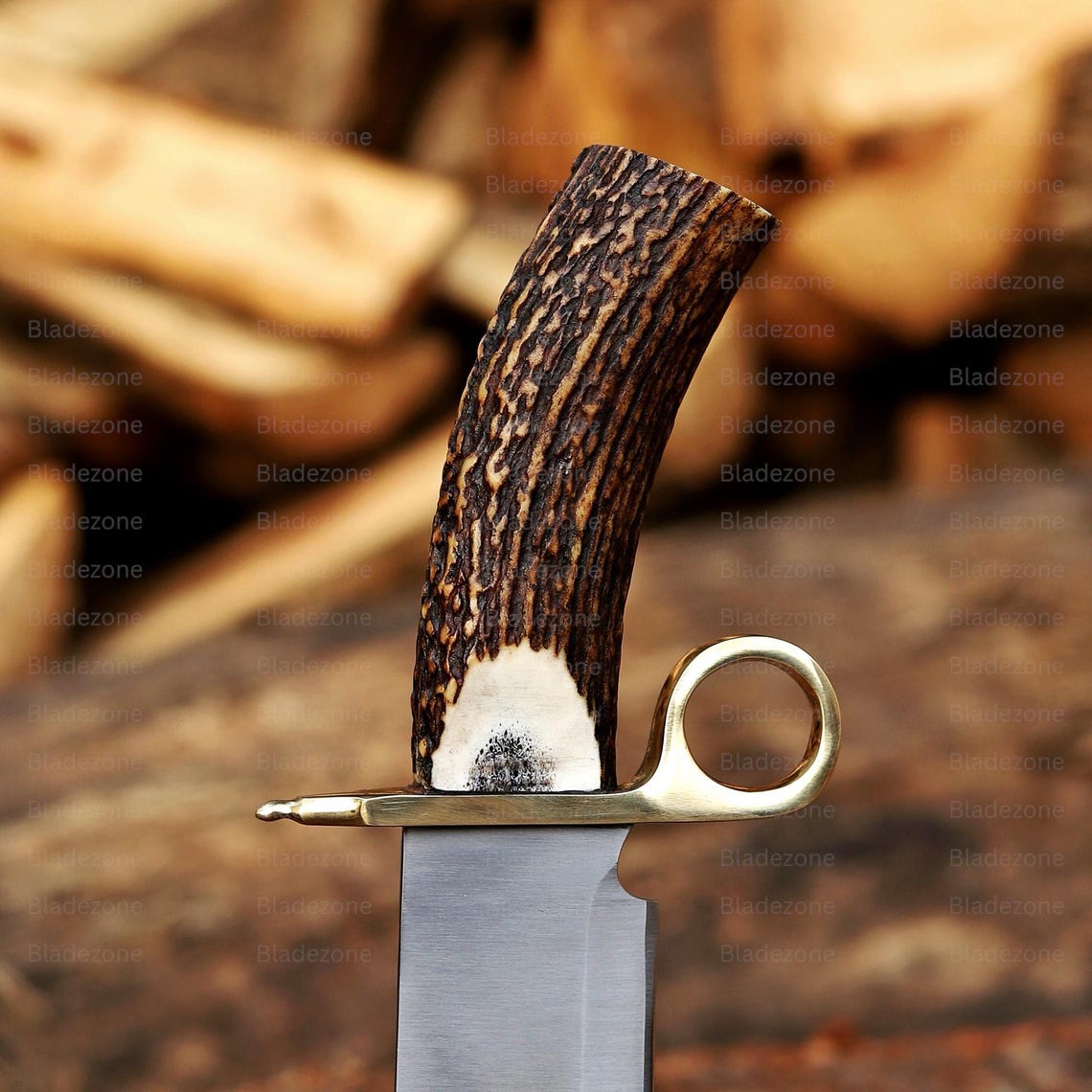 This close-up image focuses on the upper section of the handmade Bowie knife, showcasing the detailed natural stag handle with visible grain and texture. The polished brass guard and ring accent provide contrast and structure. The blade’s upper portion shows clean workmanship. The background of stacked wood enhances the knife’s rugged, outdoors-inspired aesthetic and premium handmade character.