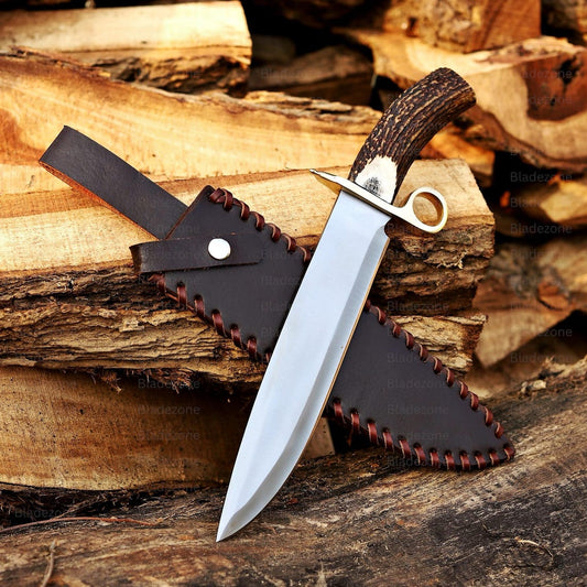 This image displays the handmade Bowie knife partially removed from its leather sheath, revealing the polished 1095 carbon steel blade. The natural stag handle and brass guard stand out with their contrasting textures. The detailed leather stitching enhances the rustic outdoor presentation, while the blade’s reflective surface highlights its craftsmanship, durability, and sharp profile suitable for hunting or collecting.