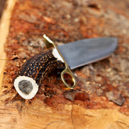 This image shows the Bowie knife being held in a gloved hand, emphasizing its size, balance, and ergonomic stag handle. The long 1095 carbon steel blade is prominently displayed, reflecting light along its polished surface. The brass ring guard enhances control and adds visual interest. The outdoor wood background reinforces the knife’s suitability for hunting, utility work, and collection.