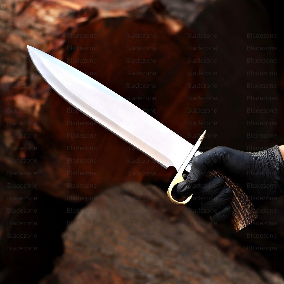 This image presents the Bowie knife firmly held in a gloved hand, highlighting the natural stag handle’s grip and the knife's impressive blade length. The polished 1095 carbon steel blade appears sharp and durable. The brass guard’s smooth curve adds elegance and strength. The rugged outdoor wood setting enhances the handcrafted and functional nature of this Bowie knife design.