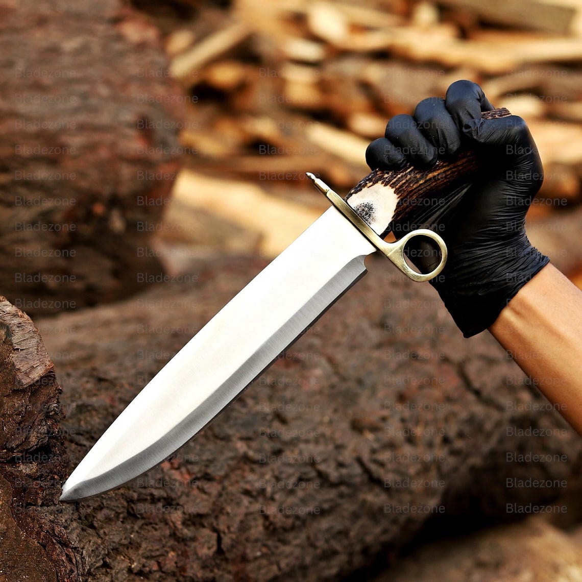 This image displays the Bowie knife standing vertically with its blade embedded in a wooden surface. The long 1095 carbon steel blade catches the light, emphasizing sharpness and polish. The natural stag handle extends upward with visible texture and color variation. The brass guard complements the knife’s overall aesthetic. The woodpile background enhances the rustic and handcrafted presentation.