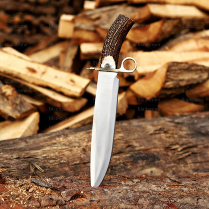 This image features a side view of the Bowie knife laid against stacked wood, highlighting the clean grind and reflective finish of the 1095 carbon steel blade. The natural stag handle arches upward, showing unique texture and pattern. The brass guard adds a classic touch to the knife’s construction. The warm wood tones reinforce the knife’s outdoor, hunting, and handcrafted qualities.