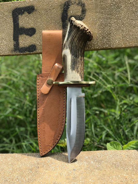 Handmade carbon steel Bowie knife featuring a real stag handle, brass guard, and high-polished blade displayed with a premium leather sheath outdoors. Perfect for hunters, collectors, or camping enthusiasts seeking a durable handmade hunting knife that combines strength, craftsmanship, and natural materials.