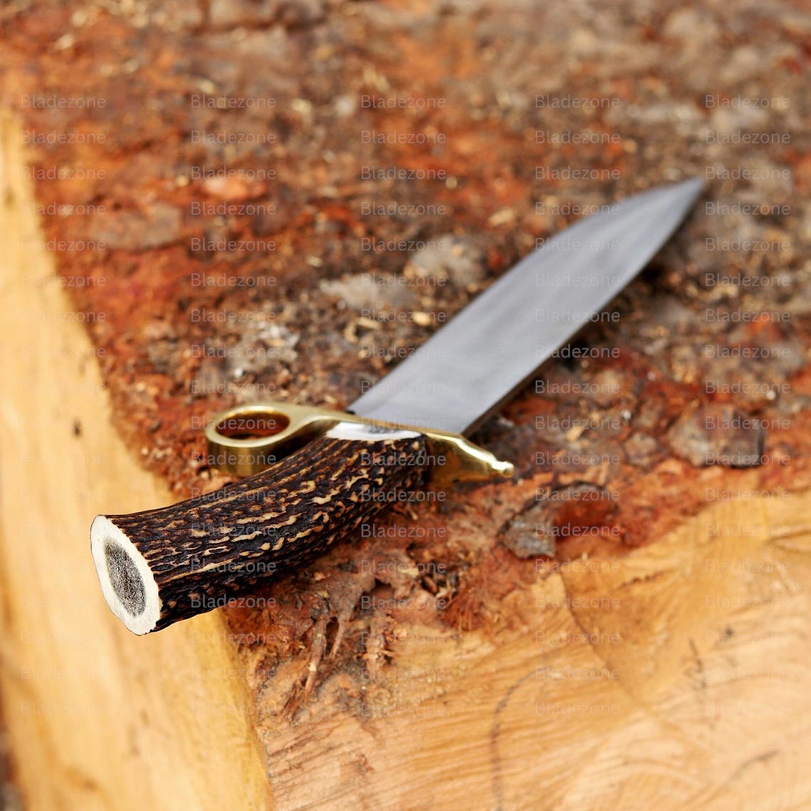 This close-up image highlights the connection between the stag handle and the brass guard of the handmade Bowie knife. The natural texture and coloration of the stag material are clearly visible, while the guard’s polished brass finish enhances the knife’s refined look. The blade’s clean grind lines emphasize craftsmanship. The background adds an earthy, outdoor atmosphere to the product’s presentation.