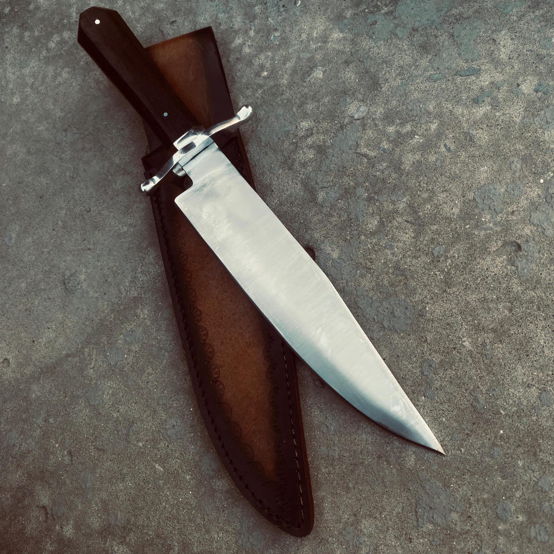 Side view of Bowie knife resting on weathered wooden block with leather sheath beside it.