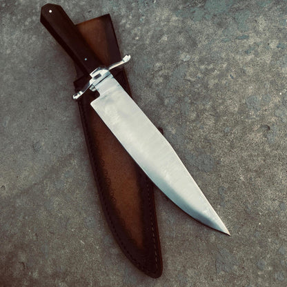 Side view of Bowie knife resting on weathered wooden block with leather sheath beside it.