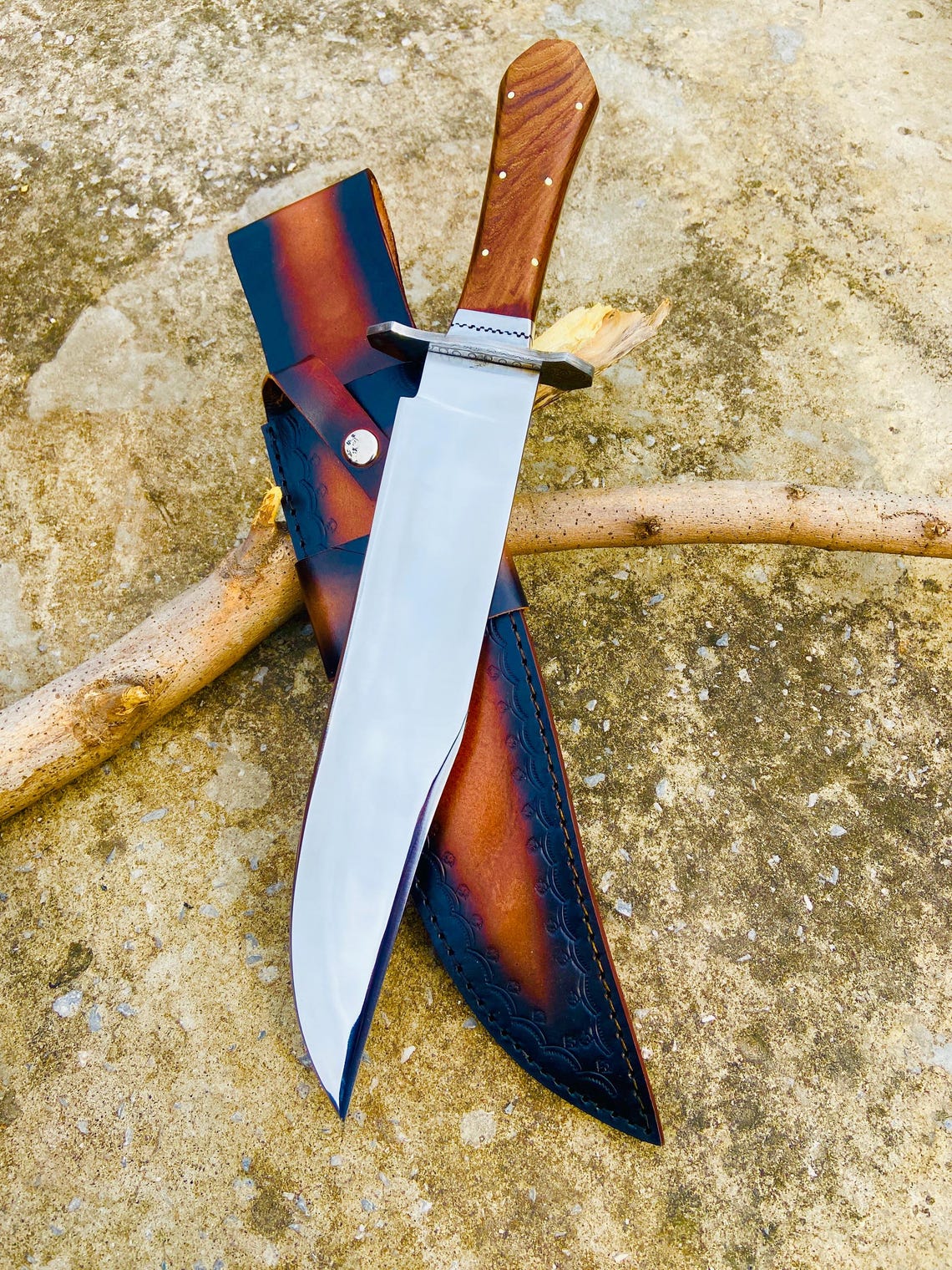 Bowie knife with mirror-polished high-carbon steel blade and rosewood coffin handle resting on a rustic wooden stick. The leather sheath shows intricate lacing and craftsmanship. A perfect handmade outdoor knife for hunting, camping, or gifting.