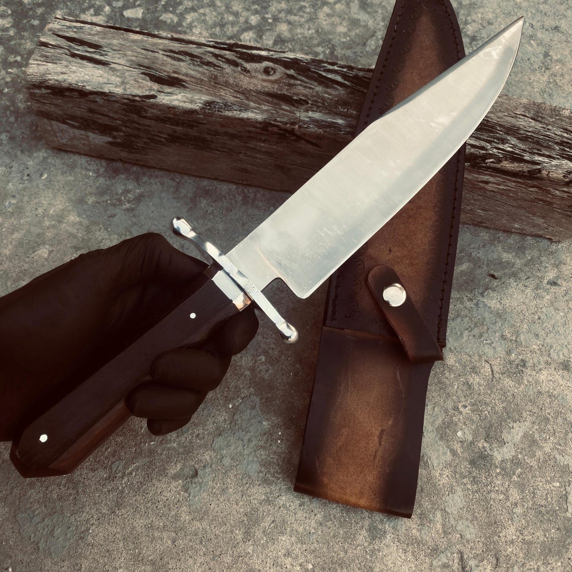 Full-length Bowie knife beside its brown hand-stitched leather sheath showing satin blade finish.