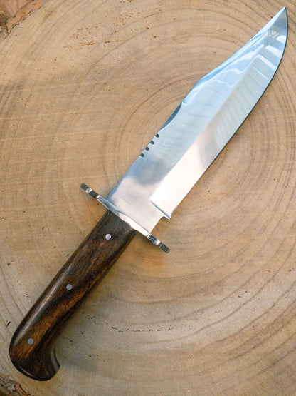 Close-up of full tang carbon steel Bowie knife showing its sharp edge, satin finish, and ergonomic wooden handle. Ideal for outdoor enthusiasts, campers, and survivalists seeking a reliable handmade blade with a premium leather sheath.