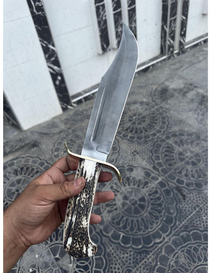 Handmade carbon steel Bowie knife with brown leather sheath displayed outdoors.