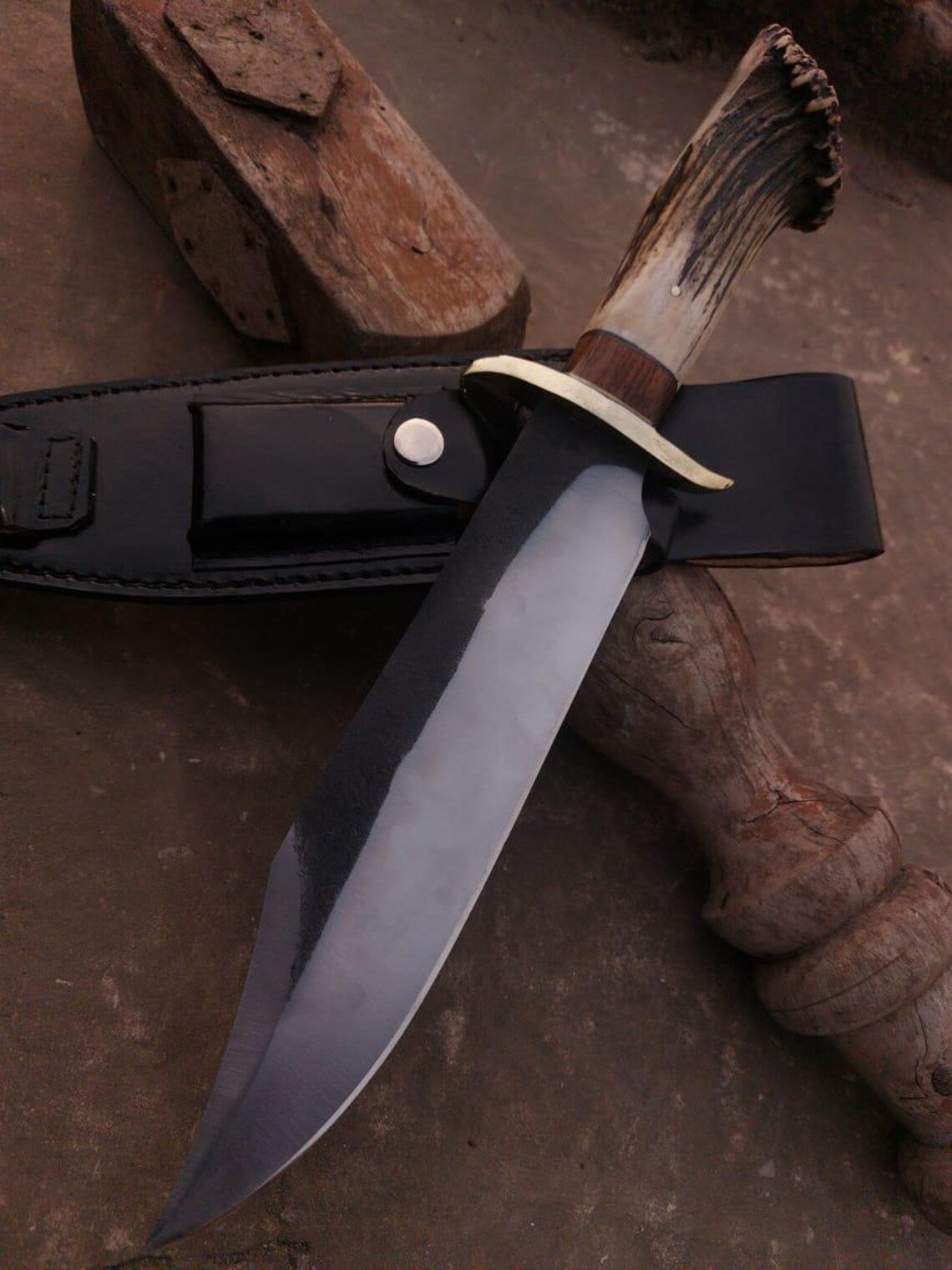 Handmade Bowie knife with polished carbon steel blade and black leather sheath on rustic surface.