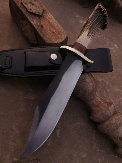 Handmade Bowie knife with polished carbon steel blade and black leather sheath on rustic surface.