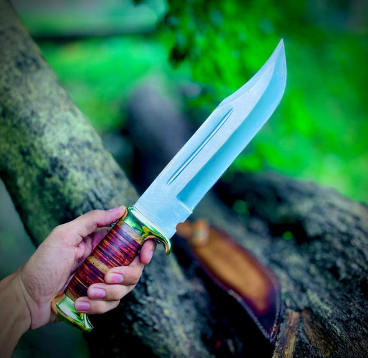 Handmade Crocodile Dundee Bowie knife featuring a sharp carbon steel blade and stacked leather handle with brass guard and pommel. The knife is displayed outdoors with its premium brown leather sheath, showcasing craftsmanship, durability, and unique design. Ideal for collectors, hunters, and outdoor adventurers looking for a reliable handmade Bowie knife with authentic styling.
