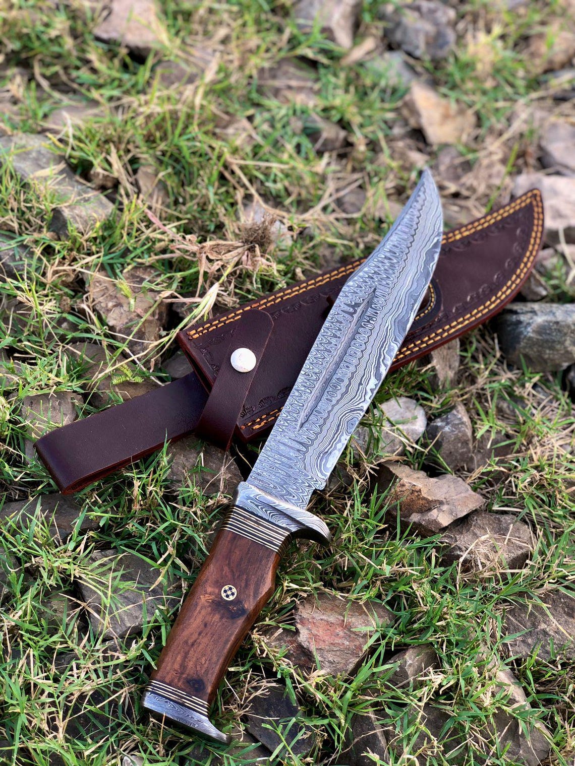 Damascus bowie knife placed on grass with sheath beside it, showing full tang design and craftsmanship.
