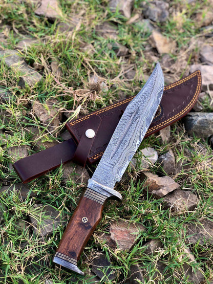 Damascus bowie knife placed on grass with sheath beside it, showing full tang design and craftsmanship.