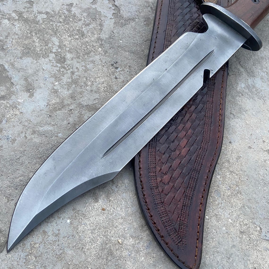 Outdoor Bowie knife with sharp clip-point carbon steel blade and hand-stitched leather sheath placed on concrete background.