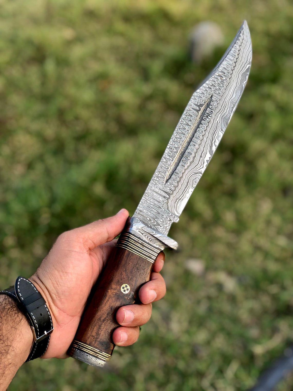 Close-up of handmade Damascus bowie knife held in hand, showing detailed blade pattern and ergonomic handle.