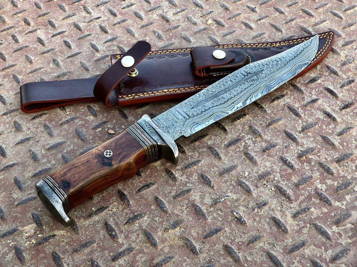 Damascus bowie knife beside its leather sheath showing blade pattern and stitching details on metal surface.