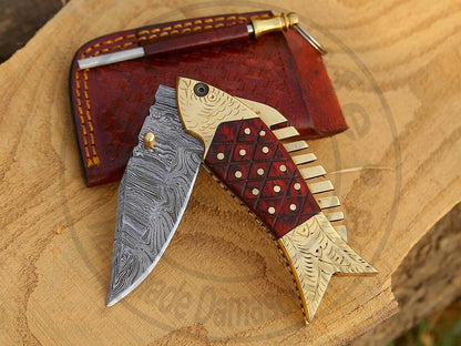 Handmade Damascus steel fish-shaped folding knife with brass and Pakka wood handle on wooden surface.