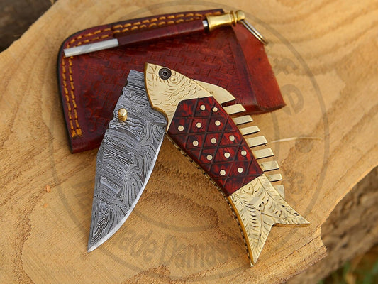 Handmade Damascus steel fish-shaped folding knife with brass and Pakka wood handle on wooden surface.