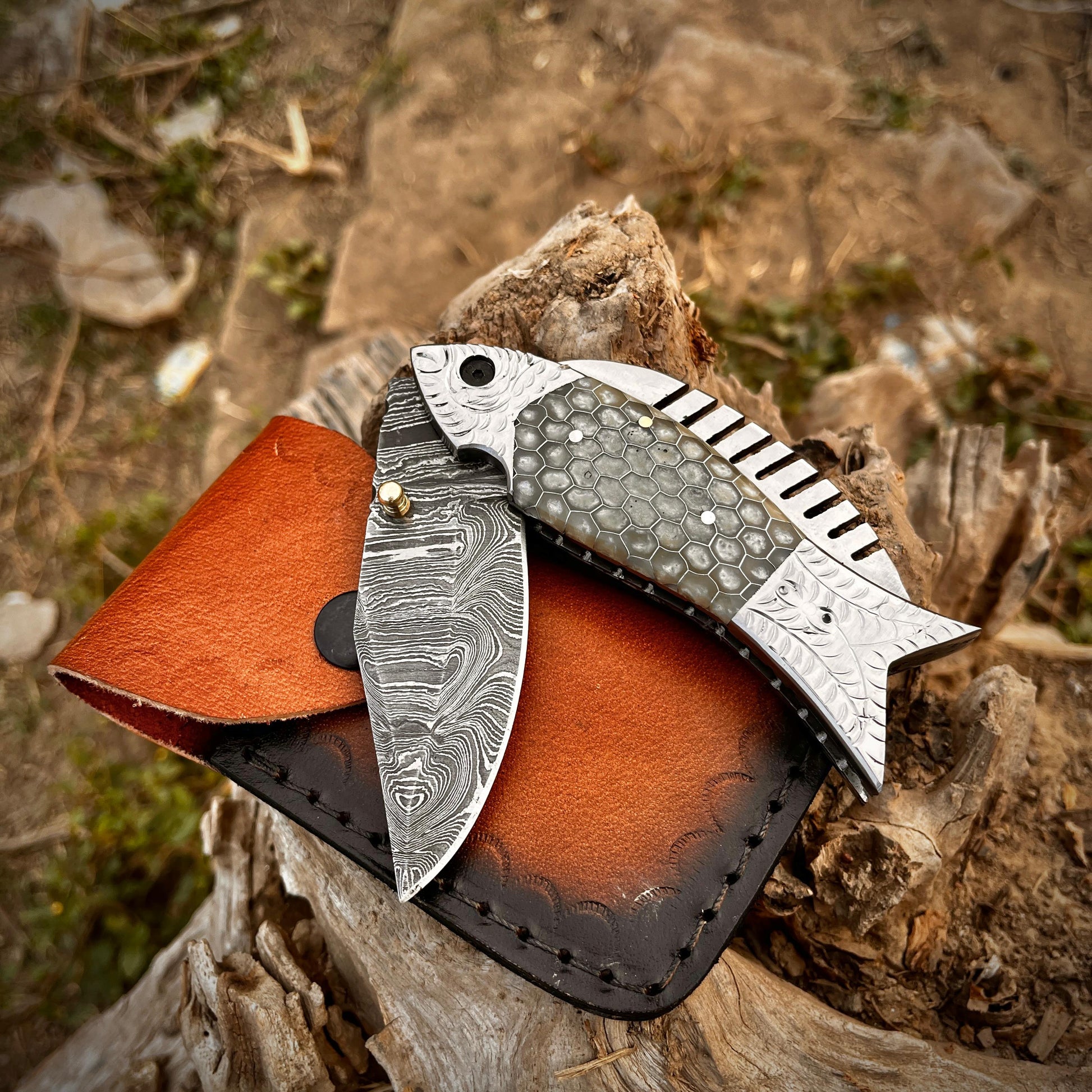 Handmade artisan fish Damascus pocket knife placed on leather sheath highlighting the unique honeycomb scale handle and folding blade design.