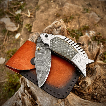 Handmade artisan fish Damascus pocket knife placed on leather sheath highlighting the unique honeycomb scale handle and folding blade design.