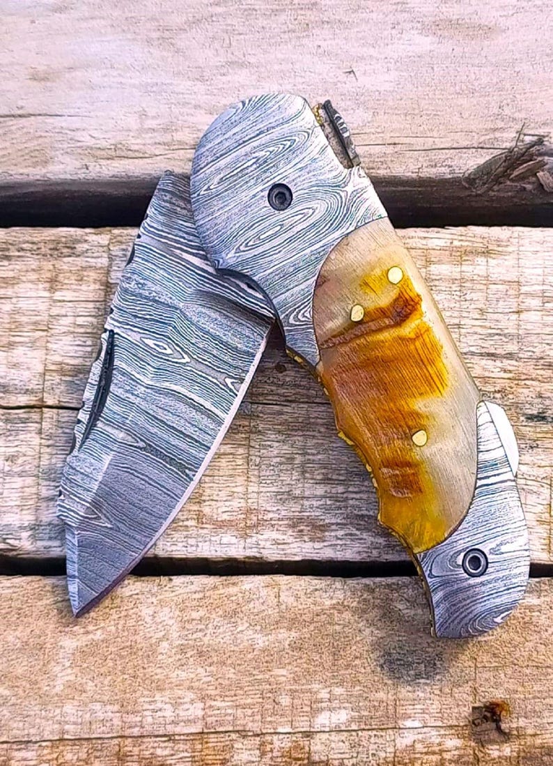 Handmade Damascus steel pocket knife with natural ram horn handle and folded blade on wood background.