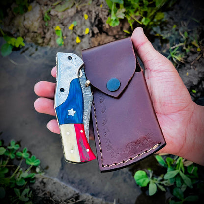 Outdoor photograph of Damascus pocket knife placed on leather pouch emphasizing everyday carry design.