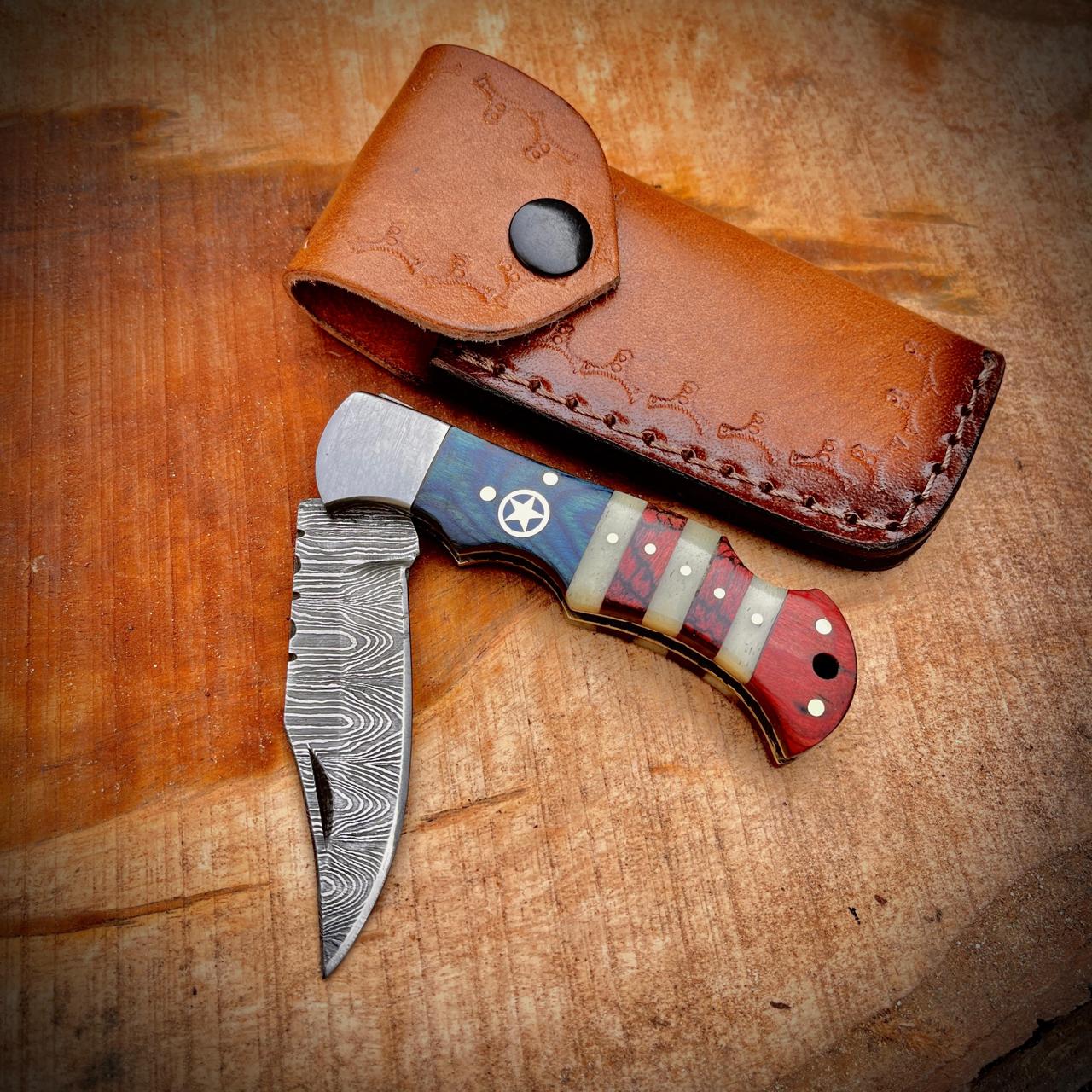 Handmade Damascus pocket knife with leather sheath displayed on a wooden tree stump in an outdoor setting showing the detailed Damascus blade pattern and unique fish shaped handle design.