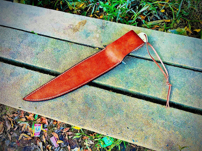 Engraved carbon steel bowie knife displayed with brown leather sheath and natural handle, outdoor background.